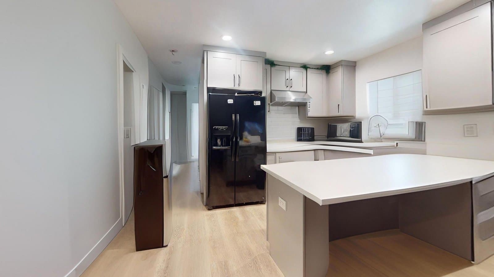 Virtual rendering: A modern kitchen with light gray cabinets, a black refrigerator, a stainless steel range hood, a white kitchen island, and light wood flooring.