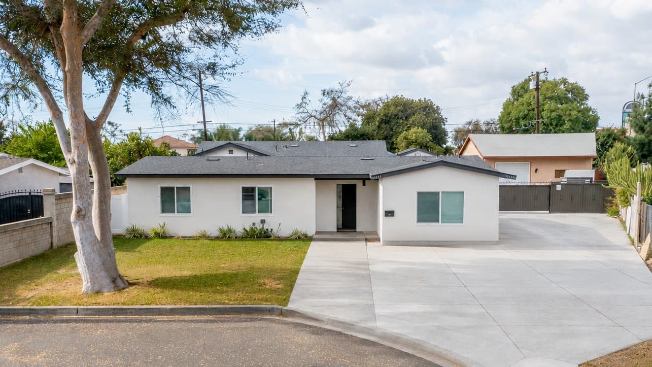 Front exterior view of a modern white house with a dark gray roof, black trim, and a black front door, identified as 13611 Glenhaven Drive. A wide concrete driveway extends along the right side leading to a gate and another building in the background. A mature tree and green lawn are in the foreground, with other houses visible beyond.